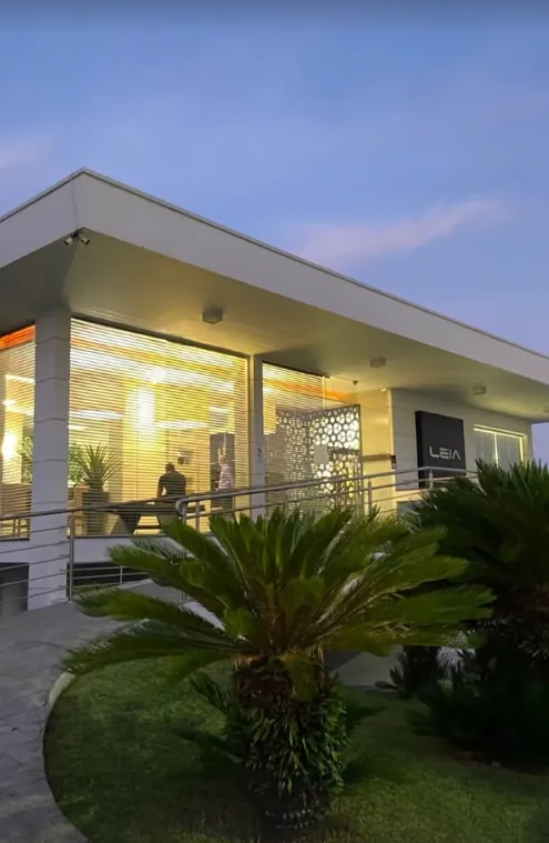Fachada da casa de Lages ao entardecer: prédio térreo branco com vidraças iluminadas, placa Leia e jardim com palmeiras