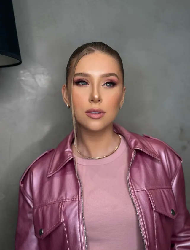 Mulher com cabelo preso liso e maquiagem rosada esfumada, de jaqueta rosa metalizada