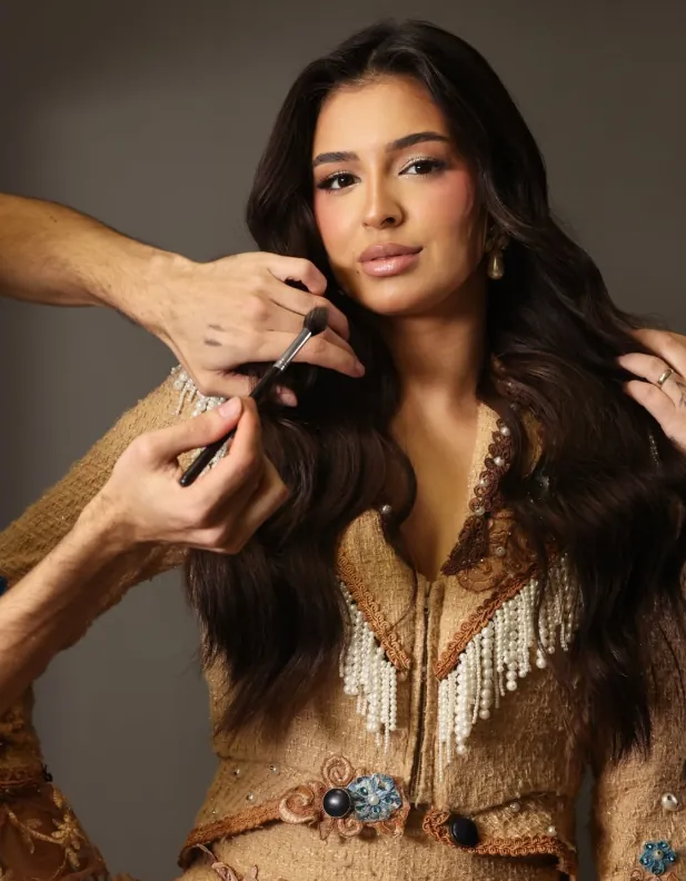 Modelo de cabelo castanho longo e ondulado sendo maquiada; mãos seguram o pincel e ajeitam os fios